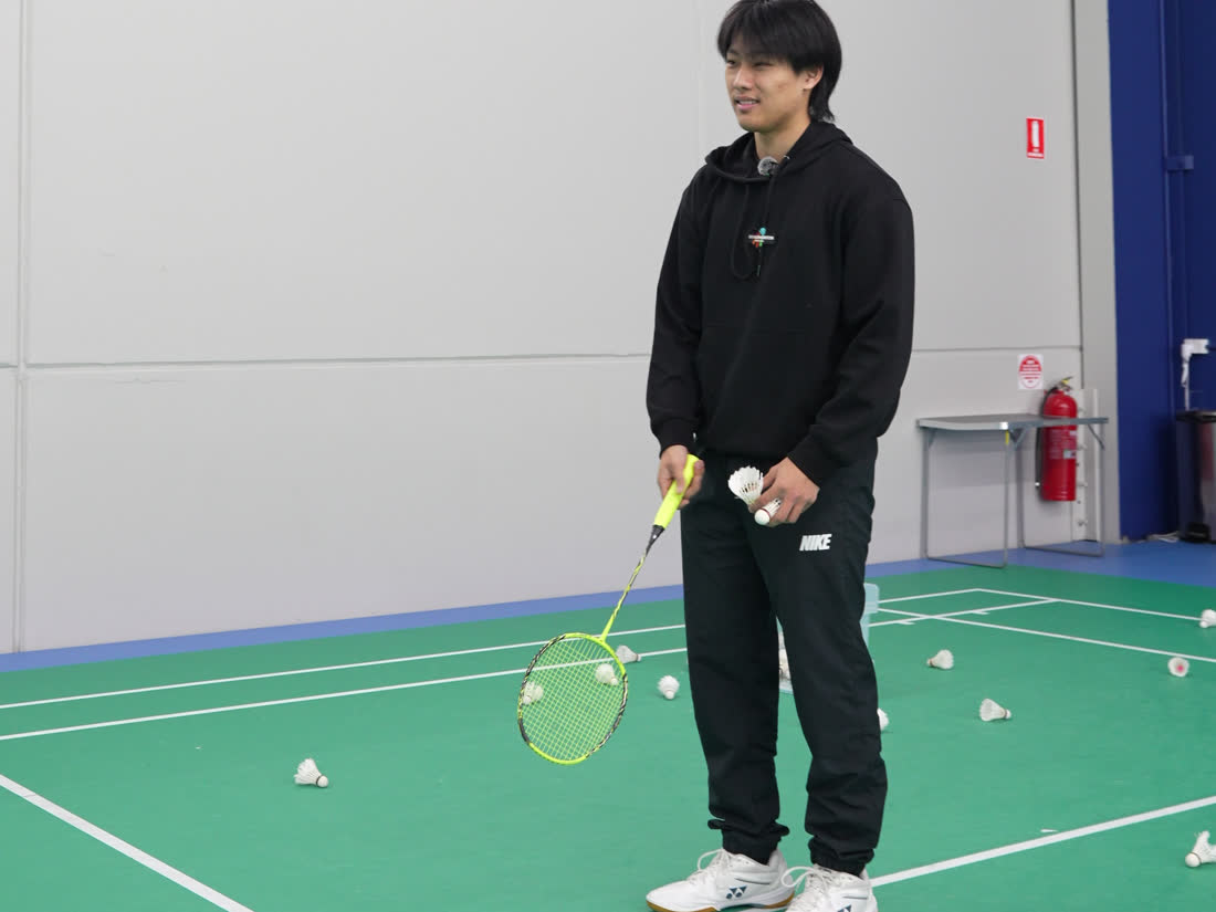 A Go Badminton coach standing on an indoor court holding a racket and a shuttle, ready to feed shuttles to a student.