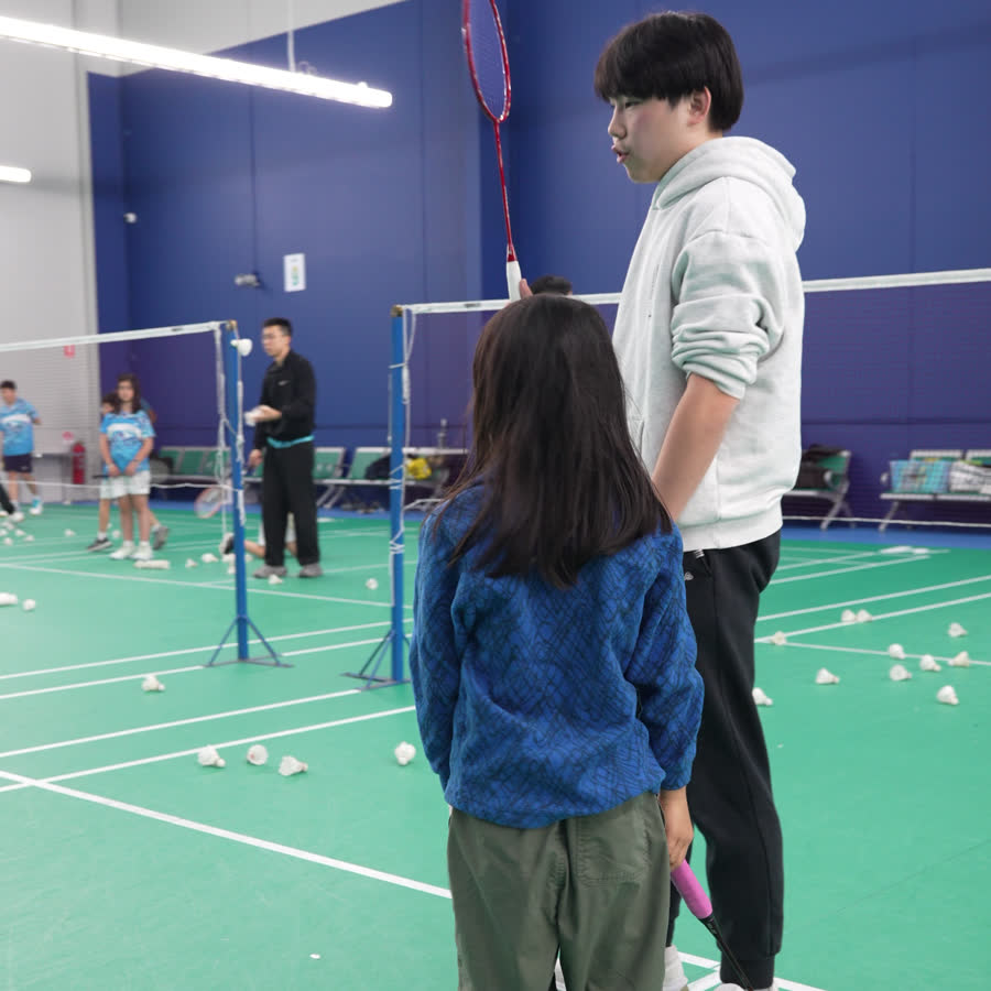A Go Badminton coach crouching beside a child on court, correcting their racquet grip.