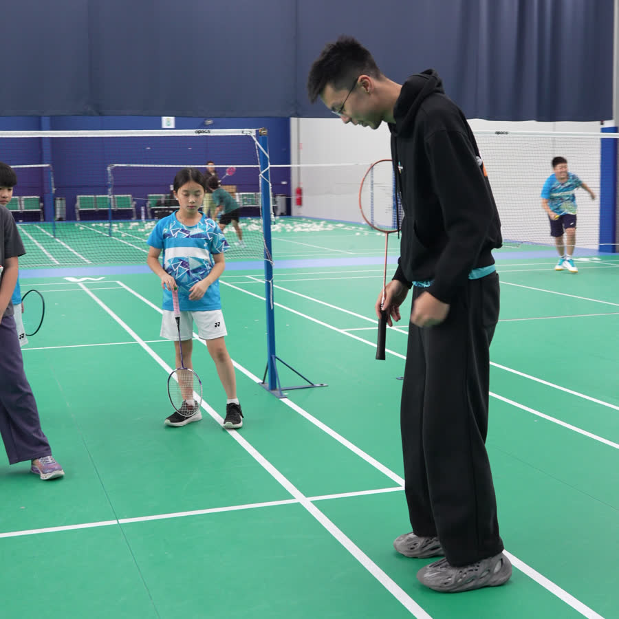 A Go Badminton coach standing on court with a small group of children holding rackets, talking them through a drill.