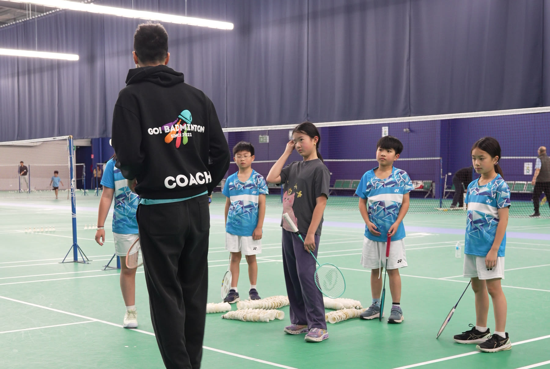 A Go Badminton coach, back to camera in a Go! Badminton coach jacket, talking to a line of five children in Go Badminton uniforms on an indoor court.